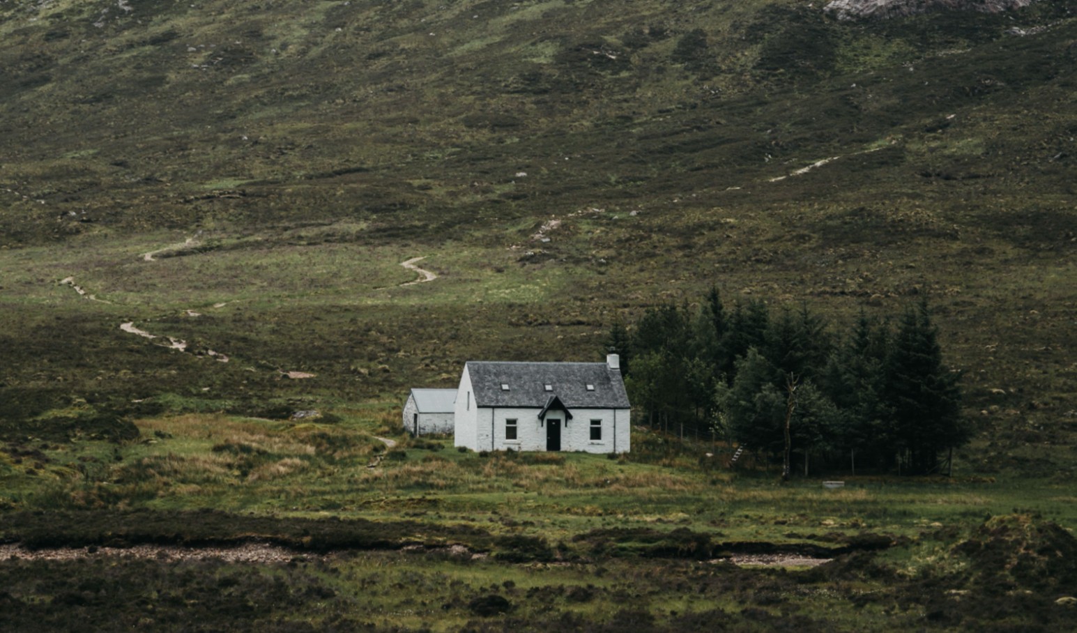 A solitary white cottage stands beneath a dramatic mountain in Glencoe, Scotland, enveloped in mist.